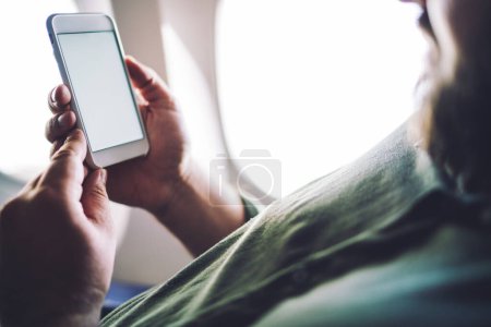 Téléchargez les photos : Vue latérale de la récolte homme barbu en vêtements décontractés assis dans l'avion près de la fenêtre et la navigation téléphone mobile pendant le vol - en image libre de droit