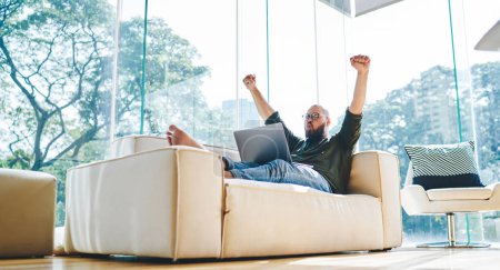 Foto de Desde abajo de hombre feliz en ropa casual sentado en el sofá en pose relajada levantando los puños mientras se celebra el logro - Imagen libre de derechos