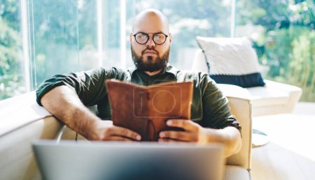 Téléchargez les photos : Jeune homme barbu concentré en tenue décontractée et lunettes de lecture livre relaxant sur un canapé confortable dans la véranda moderne par temps ensoleillé - en image libre de droit