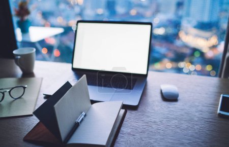 Grand angle d'ordinateur portable avec écran vierge et bloc-notes ouvert placé sur une table en bois avec des lunettes et une tasse à café près d'une fenêtre en verre dans un bureau moderne