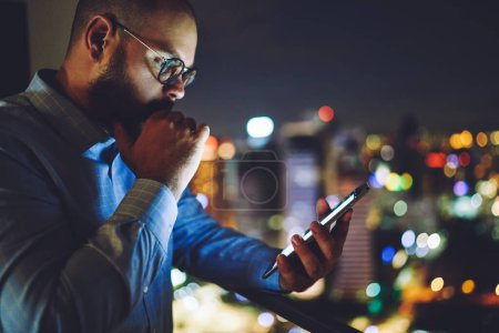 Téléchargez les photos : Vue latérale de l'entrepreneur masculin barbu de récolte debout près de la fenêtre sur fond flou de la ville moderne lèvres touchantes surf smartphone - en image libre de droit