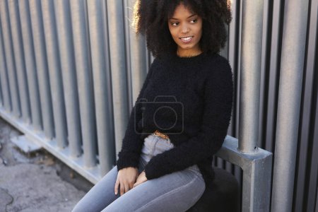 Téléchargez les photos : Cultiver jeune femme heureuse en tenue décontractée avec afro coiffure souriant et détournant les yeux tout en s'appuyant sur la clôture dans la rue - en image libre de droit