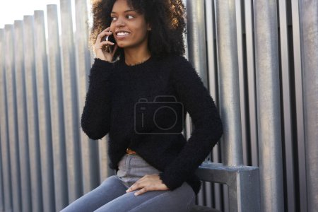 Foto de Alegre mujer afroamericana en traje casual sonriendo y mirando hacia otro lado mientras habla por teléfono móvil y se sienta en el banco - Imagen libre de derechos