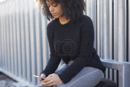 Téléchargez les photos : Femme afro-américaine pensive en tenue décontractée assise sur un banc en métal et messagerie sur un téléphone portable tout en se reposant pendant la marche - en image libre de droit