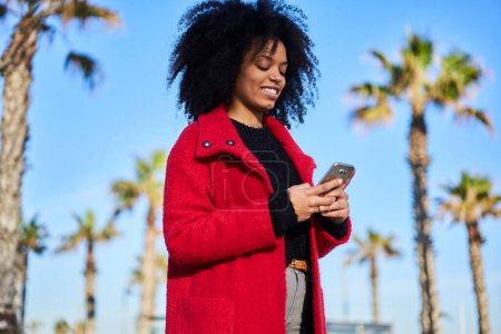 Téléchargez les photos : Faible angle de contenu Afro-Américaine en tenue décontractée souriant et bavardant sur téléphone portable tout en marchant dans la rue - en image libre de droit