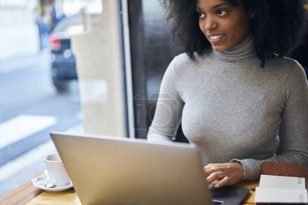 Téléchargez les photos : Jeune pigiste afro-américaine en tenue décontractée assise à table dans un espace de travail moderne et regardant loin rêveur tout en naviguant sur Internet sur netbook pendant la pause café - en image libre de droit