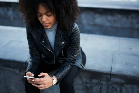 Téléchargez les photos : Pensive jeune femme afro-américaine avec les cheveux pleins d'entrain en manteau noir à la recherche d'informations sur Internet tout en se refroidissant sur le trottoir en ville - en image libre de droit