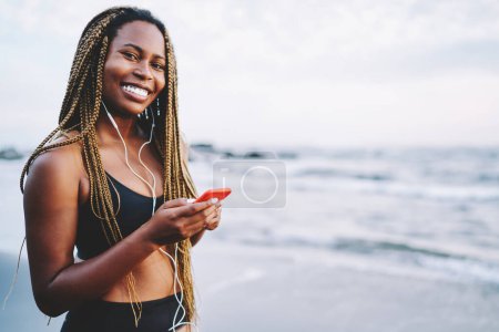 Foto de Alegre negro jogger vestida con ropa deportiva escuchar música en los auriculares conectados al teléfono en la costa, feliz mujer afroamericana con rastas utilizando aplicación de medios móviles - Imagen libre de derechos