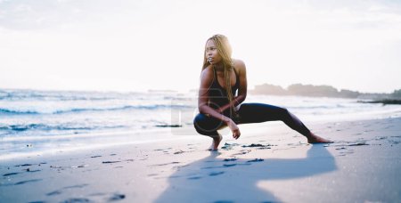 Téléchargez les photos : Flexible fit girl étirement des muscles pendant la journée de jambes passer le matin temps pour l'exercice à la plage côtière, afro-américain pieds nus joggeuse en vêtements de sport corps d'entraînement - en image libre de droit
