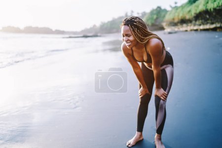 Téléchargez les photos : Joggeuse sportive afro-américaine habillée en survêtement profitant d'une pause le long du littoral le matin, joyeuse femelle à la peau foncée se réjouissant et riant après l'entraînement sportif sur la côte - en image libre de droit