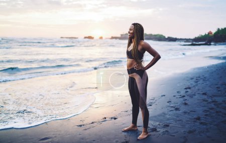 Foto de Alegre mujer afroamericana sintiendo felicidad durante el entrenamiento matutino en la costa mirando hacia otro lado y sonriendo, divertido corredor de piel oscura vestido con ropa deportiva riendo a la orilla del mar - Imagen libre de derechos