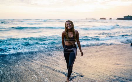Foto de Retrato de chica afroamericana en forma despreocupada en chándal sonriendo a la cámara durante la mañana en la orilla del mar, corredor de piel oscura feliz en top deportivo disfrutando de un estilo de vida saludable y positivo para el cuerpo - Imagen libre de derechos