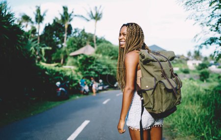 Téléchargez les photos : Joyeuse touriste afro-américaine avec sac à dos touristique souriant pendant les vacances d'été pour visiter l'île tropicale, heureuse femme à la peau foncée avec sac à dos de voyage explorant l'environnement - en image libre de droit