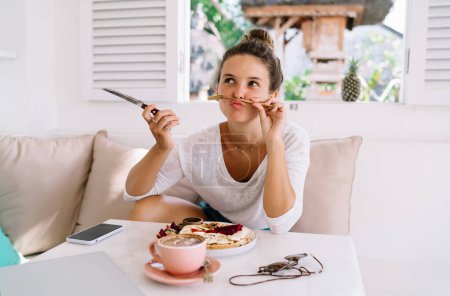 Téléchargez les photos : Charmante femme en vêtements de nuit assise à table avec une tasse de café et un délicieux repas tout en s'amusant pendant le petit déjeuner le matin - en image libre de droit