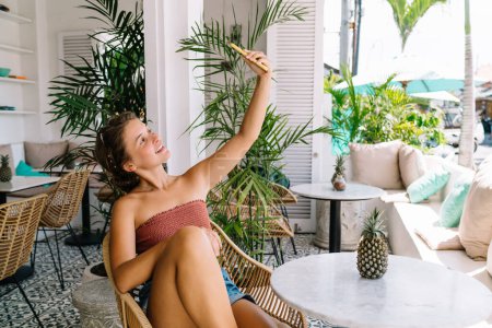 Téléchargez les photos : Touriste féminine joyeuse dans des vêtements élégants assis sur une chaise en osier près de la table ronde et prenant selfie sur smartphone dans une chambre spacieuse - en image libre de droit