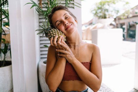 Téléchargez les photos : Femme aux cheveux bruns souriante en vêtements d'été assise sur une chaise et embrassant l'ananas frais pendant ses vacances dans un pays tropical - en image libre de droit