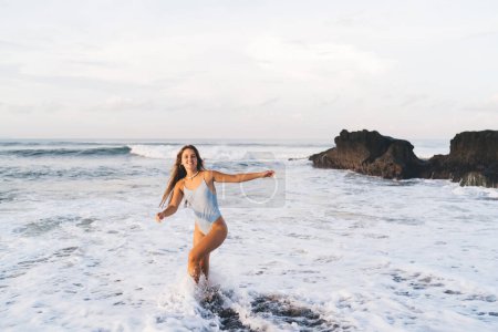 Téléchargez les photos : Contenu complet du corps jolie femme portant des maillots de bain blancs marchant sur le bord de la mer lavé par agitant la mer mousseuse par beau temps et en regardant la caméra - en image libre de droit