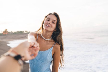 Téléchargez les photos : Jeune jolie femelle portant maillot de bain bleu tenant les copains main doucement tout en se tenant sur le bord de mer humide sablonneux et en regardant la caméra - en image libre de droit