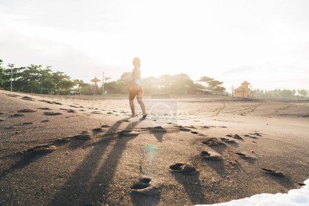 Téléchargez les photos : Vue arrière de la femme heureuse en maillots de bain et lunettes de soleil jogging flirt sur la rive sablonneuse humide de Bali exotique au coucher du soleil lumineux retour éclairé - en image libre de droit