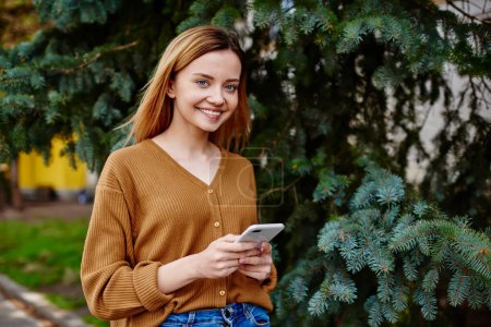 Foto de Mujer joven feliz en ropa casual mirando a la cámara mientras mensajes de texto en el teléfono inteligente de pie cerca de los árboles de coníferas en el parque - Imagen libre de derechos