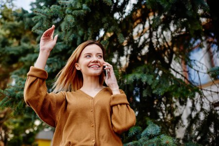 Foto de Desde abajo de la joven mujer encantada en traje casual hablando por teléfono con un amigo saludando de la mano y mirando hacia otro lado contra las ramas de árbol de coníferas en el parque - Imagen libre de derechos