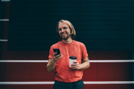 Téléchargez les photos : Jeune homme moderne barbu souriant en t-shirt rouge textant sur téléphone portable tenant tasse de café à emporter dans la rue de la ville - en image libre de droit