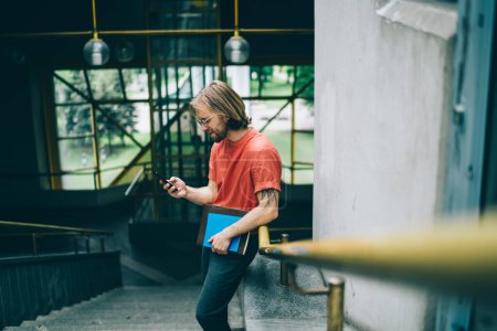 Foto de Hombre joven barbudo enfocado en gafas con pelo largo en mensajería de camiseta roja en el teléfono móvil sosteniendo carpetas en la mano en la construcción - Imagen libre de derechos