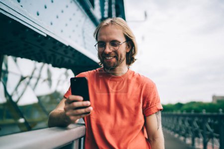 Téléchargez les photos : Contenu homme barbu rouge souriant dans des lunettes avec les cheveux longs en t-shirt rouge textos sur téléphone portable debout sur le pont - en image libre de droit
