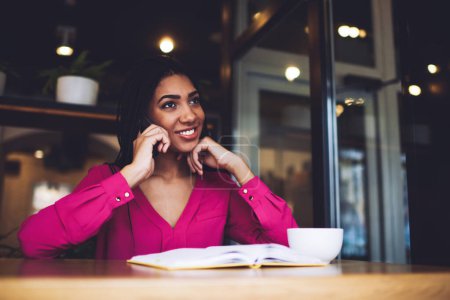 Téléchargez les photos : Angle bas de femme afro-américaine joyeuse en vêtements décontractés roses avec sourire et la main à menton regardant loin et parlant sur téléphone mobile - en image libre de droit