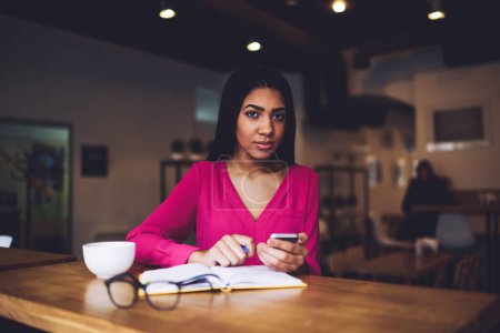 Téléchargez les photos : Femme noire réfléchie en tenue décontractée lumineuse regardant la caméra assise à la cafétéria avec téléphone mobile et vérifiant les réseaux sociaux - en image libre de droit