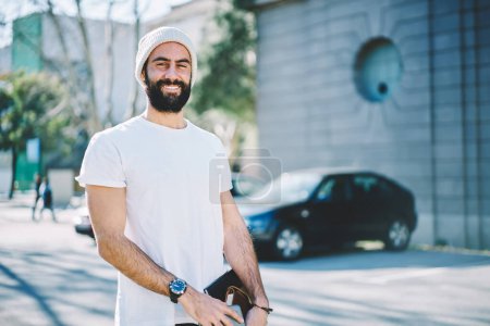 Half length portrait of cheerful Turkish student in hat smiling at camera holding education equipment in hand, happy Middle Eastern male with textbooks posing at street urban setting on leisure