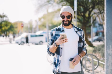 Glückliche männliche Blogger aus dem Nahen Osten in den 30er Jahren, die während ihres Urlaubs und zur Kaffeezeit in der Stadt Netznachrichten checken, türkischer Hipster mit Sonnenbrille und Hut, der Tickets für touristische Urlaube bucht