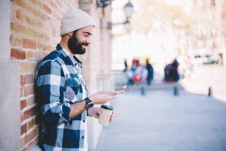 Glückliche männliche Blogger aus dem Nahen Osten in den 30er Jahren beim Überprüfen von Netzwerknachrichten während des Urlaubs und zur Kaffeezeit in der Stadt, Seitenansicht eines türkischen Hipsters mit Hut, der Tickets für touristische Urlaube bucht