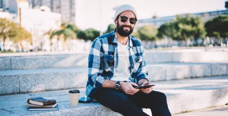 Portrait of trendy Middle Eastern male blogger using palmtop technology during solo travel vacations, Turkish hipster guy in sunglasses holding digital tablet and looking at camera during free time