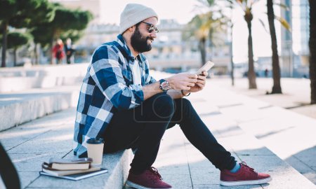 Trendy gekleidete männliche Touristen surfen auf Webseiten, um Reiseberichte zu lesen, die sich bei Coffee to go und Bildungsausrüstung an städtischen Treppen ausruhen.