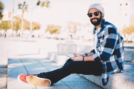 Retrato del guapo blogger turco en auriculares electrónicos mirando a la cámara durante el ocio en la ciudad, tipo hipster barbudo en gafas de sol y auriculares escuchando música lista de reproducción a través de la aplicación móvil de medios