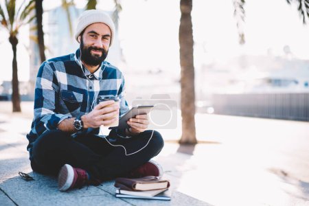 Retrato de estudiante turco con café para ir a escuchar audio libro de educación y mirando a la cámara descansando en la ciudad, chico hipster de Oriente Medio en auriculares electrónicos usando el panel táctil