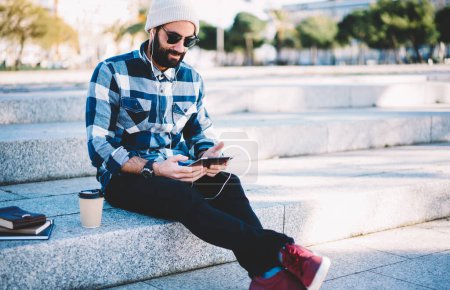 Bearded male blogger dressed in stylish clothing spending leisure time at urban setting listening online podcast, Turkish hipster guy in earphones and sunglasses watching video vlog via touch pad