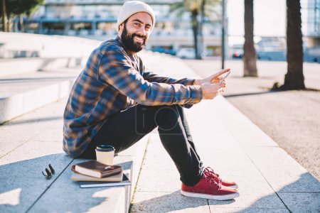 Portrait of happy bearded hipster guy in stylish clothes smiling at camera sitting at urban stairs with notepads and coffee to go enjoying leisure time outdoors, Middle Eastern blogger with smartphone