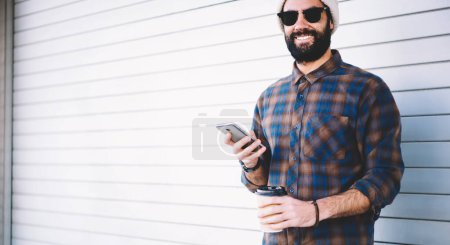 Portrait à mi-longueur du gars hipster joyeux tenant la technologie mobile et à emporter boisson caféine et souriant à la caméra pendant le voyage de vacances, heureux blogueur masculin du Moyen-Orient avec cellulaire