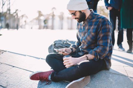 Middle Eastern backpacker typing text of travel publication for sharing to social networks during journey trip, bearded tourist checking received messages on smartphone enjoy coffee time in city