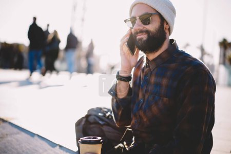 Millennial hipster chico en gafas de sol tienen conversación de llamada durante las vacaciones internacionales, Oriente Medio masculina celebración de bebidas con cafeína haciendo itinerancia teléfono inteligente de comunicación descansando en la ciudad