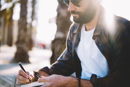 Hinterleuchteter Blick auf einen männlichen Autor aus dem Nahen Osten, der Textideen in einen Notizblock schreibt, kreativer Hipster mit Sonnenbrille und Hemd, der informative Notizen macht, während er touristische Besichtigungen plant
