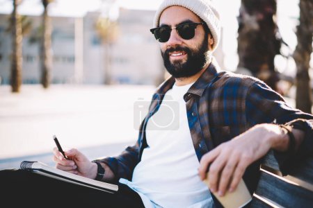 Porträt eines trendig gekleideten männlichen Studenten mit Bart, der in die Kamera schaut, während er auf einer städtischen Bank in der Stadt lernt, Schriftsteller aus dem Nahen Osten mit trendiger Sonnenbrille, der Reisezeit für die Reiseplanung verbringt