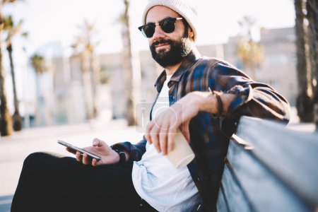 Portrait d'un blogueur turc barbu avec téléphone portable et tasse à emporter dans les mains regardant la caméra, homme du Moyen-Orient en lunettes de soleil en utilisant la technologie smartphone pendant le temps du café en plein air
