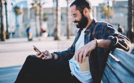 Bärtiger Tourist mit koffeinhaltigem Getränk mit Handy-Anwendung zum Lesen von Publikationstexten, der sich auf einer Bank ausruht, männlicher Blogger aus dem Nahen Osten mit Sonnenbrille, der Medieninhalte per Mobiltelefon durchsucht