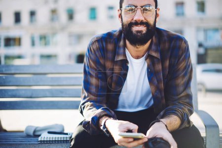 Portrait of happy Turkish blogger in stylish sunglasses using mobile technology during free time in city, cheerful hipster guy with beard holding modern cellular device and smiling at camera