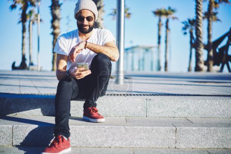 Nahost-Mann mit trendiger Sonnenbrille liest SMS auf Mobiltelefonen, bärtiger Hipster telefoniert und chattet mit einer Anwendung zur Online-Buchung im Internet
