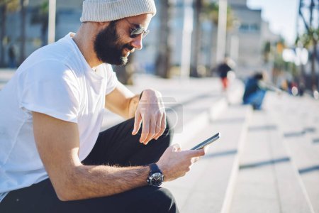 Hombre de Oriente Medio en gafas de sol de moda lectura de mensaje de texto recibido en la tecnología celular, barbudo hipster chico de telefonía y chat móvil utilizando la aplicación para hacer la reserva en línea web