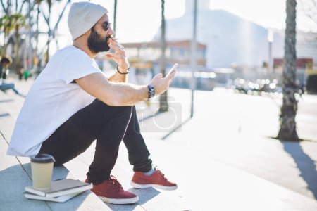 Moderno hombre vestido de turismo fumar cigarrillo y mensajería durante las vacaciones de conexión a 4g para el envío de SMS de texto, hombre turco en gafas de sol y sombrero de lectura de la publicación móvil de navegación página inalámbrica